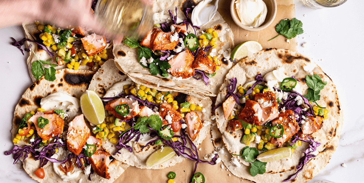 Hand assembling colourful cajun salmon tacos, reaching for toppings including corn, feta, time and avocado.