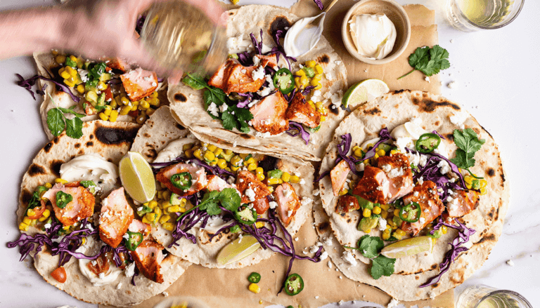 Hand assembling colourful cajun salmon tacos, reaching for toppings including corn, feta, time and avocado.